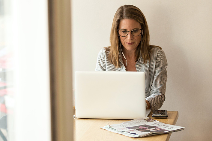 A person works on a laptop with a newspaper on the table.