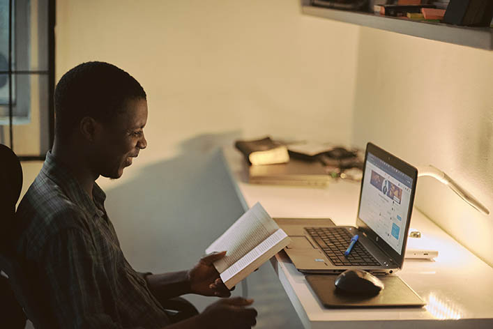 Man reads a book while sitting at a laptop.